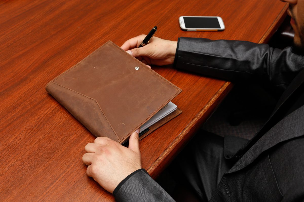 Close up of someone sitting at a wood table with a leather folio and smartphone.