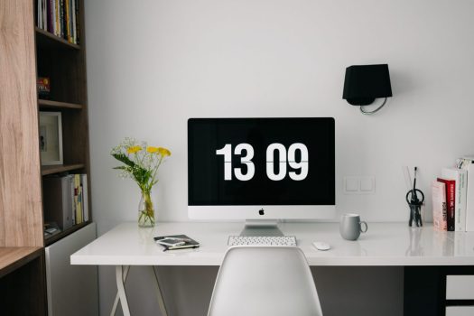 a home office desk with a large desktop screen displaying a clock and a vase with yellow flowers beside a bookcase