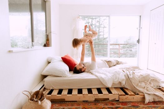 a father and child playing on a bed in a brightly lit bedroom with white walls and a wooden pallet bedframe