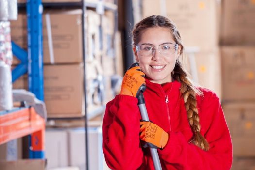 a woman wearing safety glasses and orange gloves smiles in a warehouse