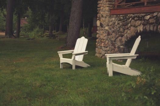 two Adirondack type white lawn chairs sit on a grassy lawn beside an old stone house