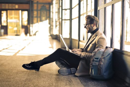 a young man in a turtleneck and glasses sits on the floor and smiles at his laptop like he is video chatting with someone