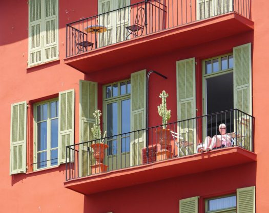 an older woman sits on her balcony beside potted cacti in a salmon colored building