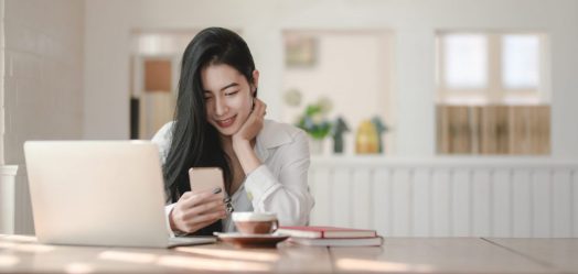 a woman with long black hair sits at a table with her laptop and coffee. she is holding her phone and smiling at it.