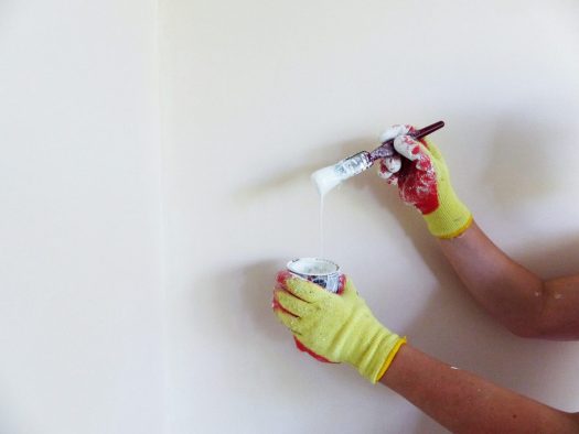 A pair of hands in yellow work gloves is holding a can of white paint and a paintbrush dripping white paint.
