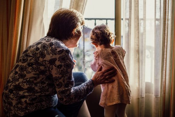 An older woman and a toddler stand together looking out a window. You can only see the backs of their heads.