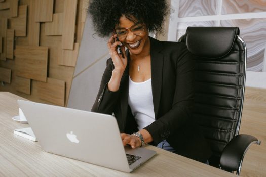 a black woman talks on the phone and smiles while working on her laptop